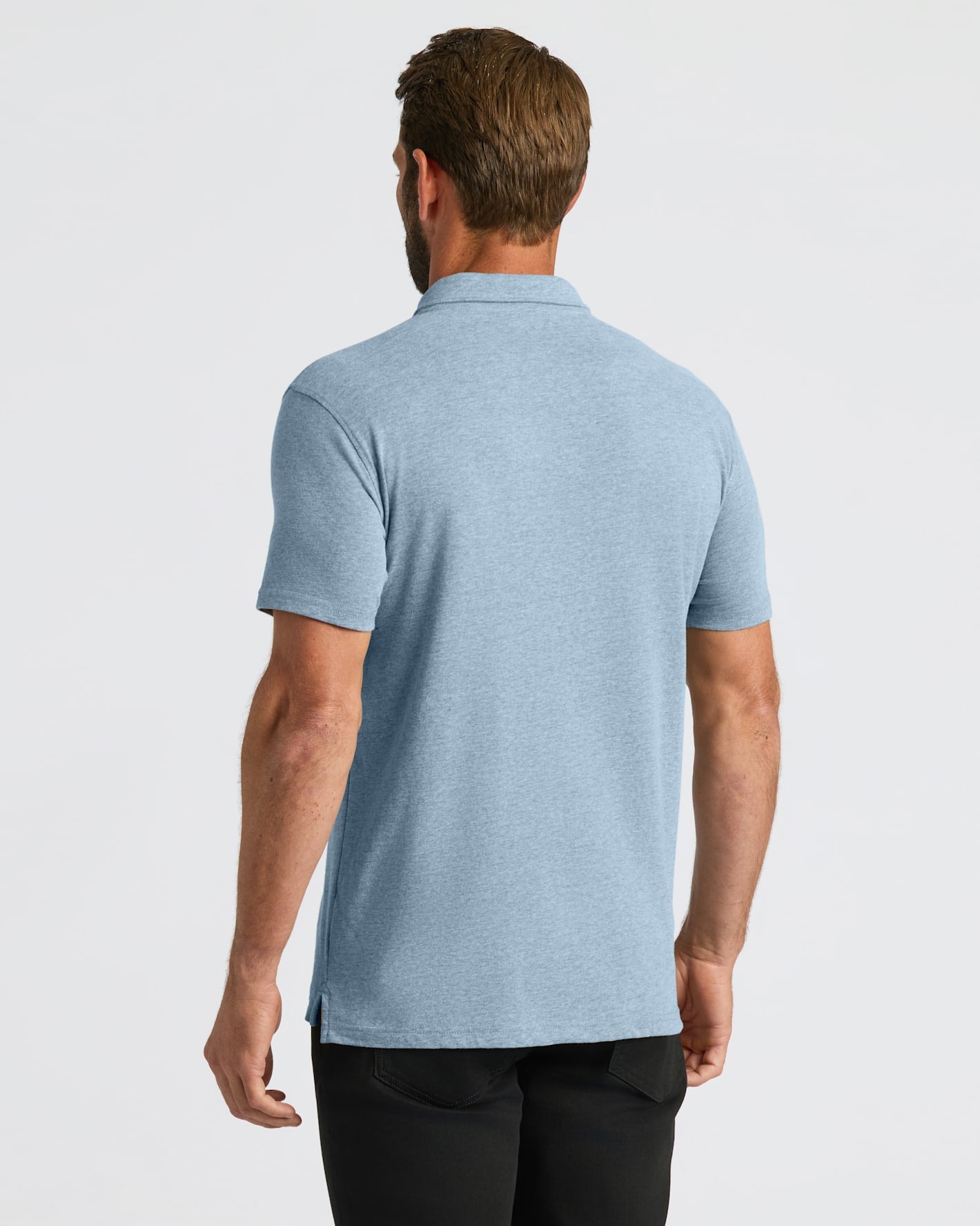 A man viewed from the back wearing a light heather blue short-sleeve polo shirt and black pants, against a plain light gray studio background.