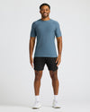 A smiling man wearing a Baltic Active Crew Neck Tee in blue paired with black shorts, white socks, and white sneakers, standing against a plain light background.