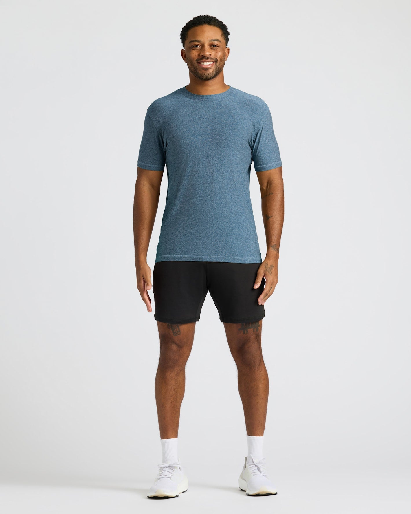 A smiling man wearing a Baltic Active Crew Neck Tee in blue paired with black shorts, white socks, and white sneakers, standing against a plain light background.