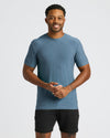 Man wearing a heather blue Baltic Active Crew Neck Tee paired with black shorts, smiling and standing against a plain white background in a casual pose.