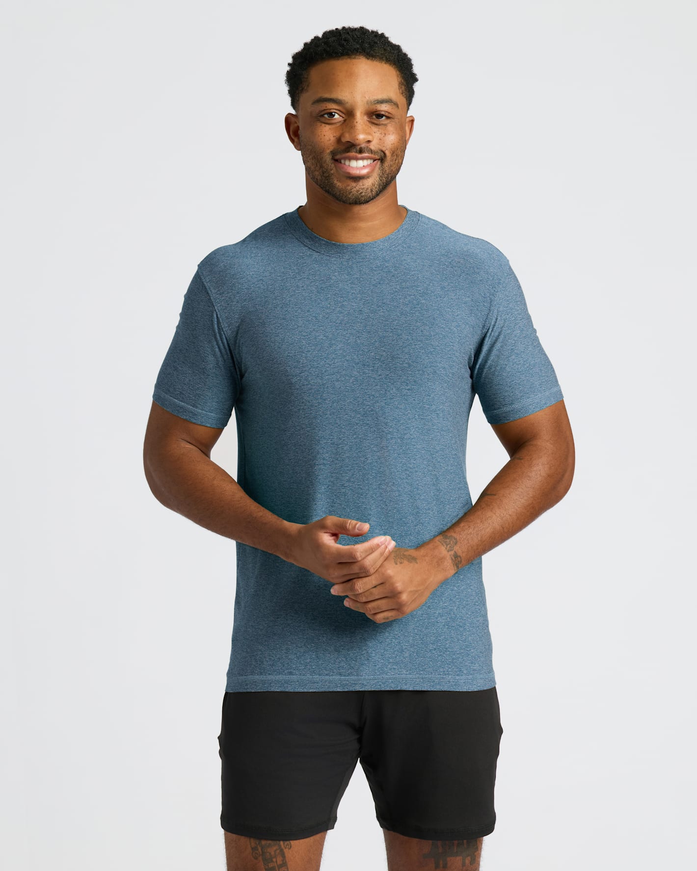 Man wearing a heather blue Baltic Active Crew Neck Tee paired with black shorts, smiling and standing against a plain white background in a casual pose.