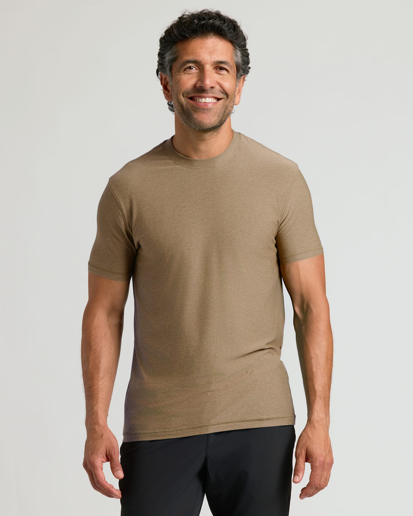 Man wearing a fitted Driftwood Active Crew Neck Tee in a neutral brown shade paired with black shorts, smiling confidently against a plain background.