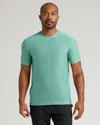 A man wearing a short-sleeve crewneck t-shirt in a solid mint green color, paired with black pants, standing against a light gray studio background.