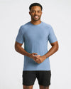 A man wearing a heathered light blue crewneck t-shirt and black athletic shorts, standing against a light gray studio background.