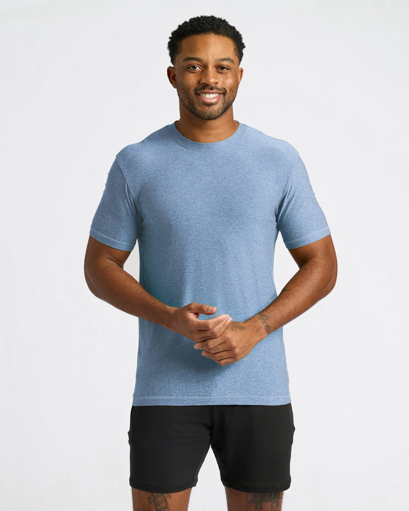 A man wearing a heathered light blue crewneck t-shirt and black athletic shorts, standing against a light gray studio background.