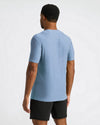 A man viewed from behind wearing a light heather blue short-sleeve t-shirt and black athletic shorts against a plain white studio background.