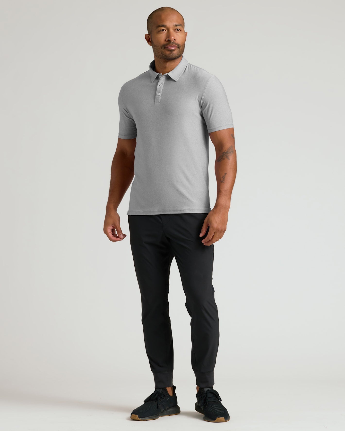 Man wearing a Light Heather Gray Active Polo 2.0 with short sleeves, paired with black pants and black sneakers, standing against a plain light background.