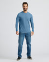 A man modeling a light blue long-sleeve crewneck t-shirt and blue jeans against a light gray studio background.