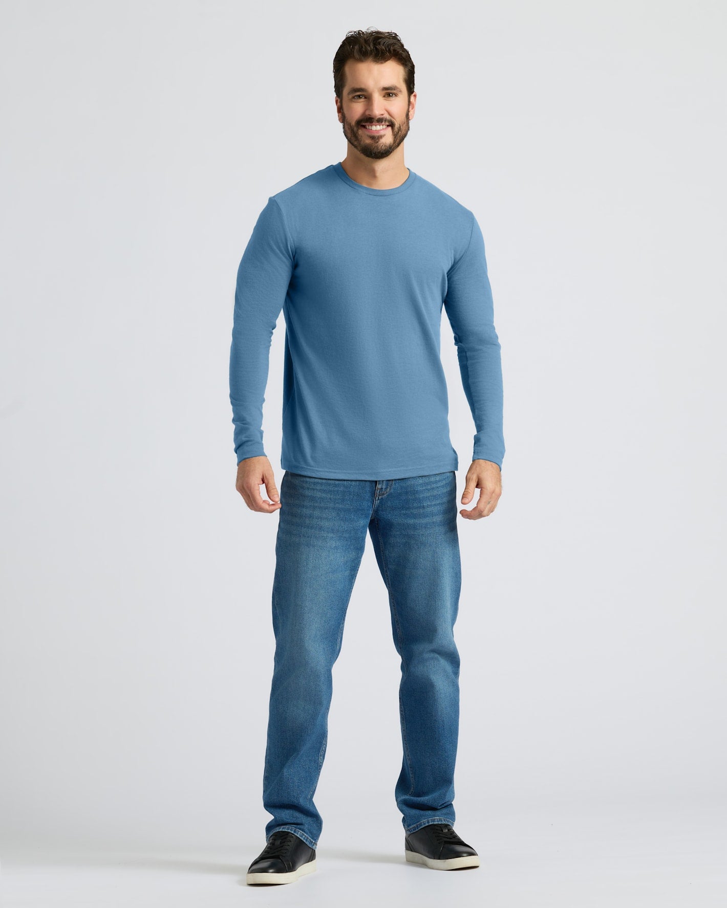 A man modeling a light blue long-sleeve crewneck t-shirt and blue jeans against a light gray studio background.