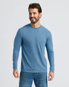 A smiling man wearing a light blue long-sleeve crewneck shirt and blue jeans, standing against a light gray studio background.