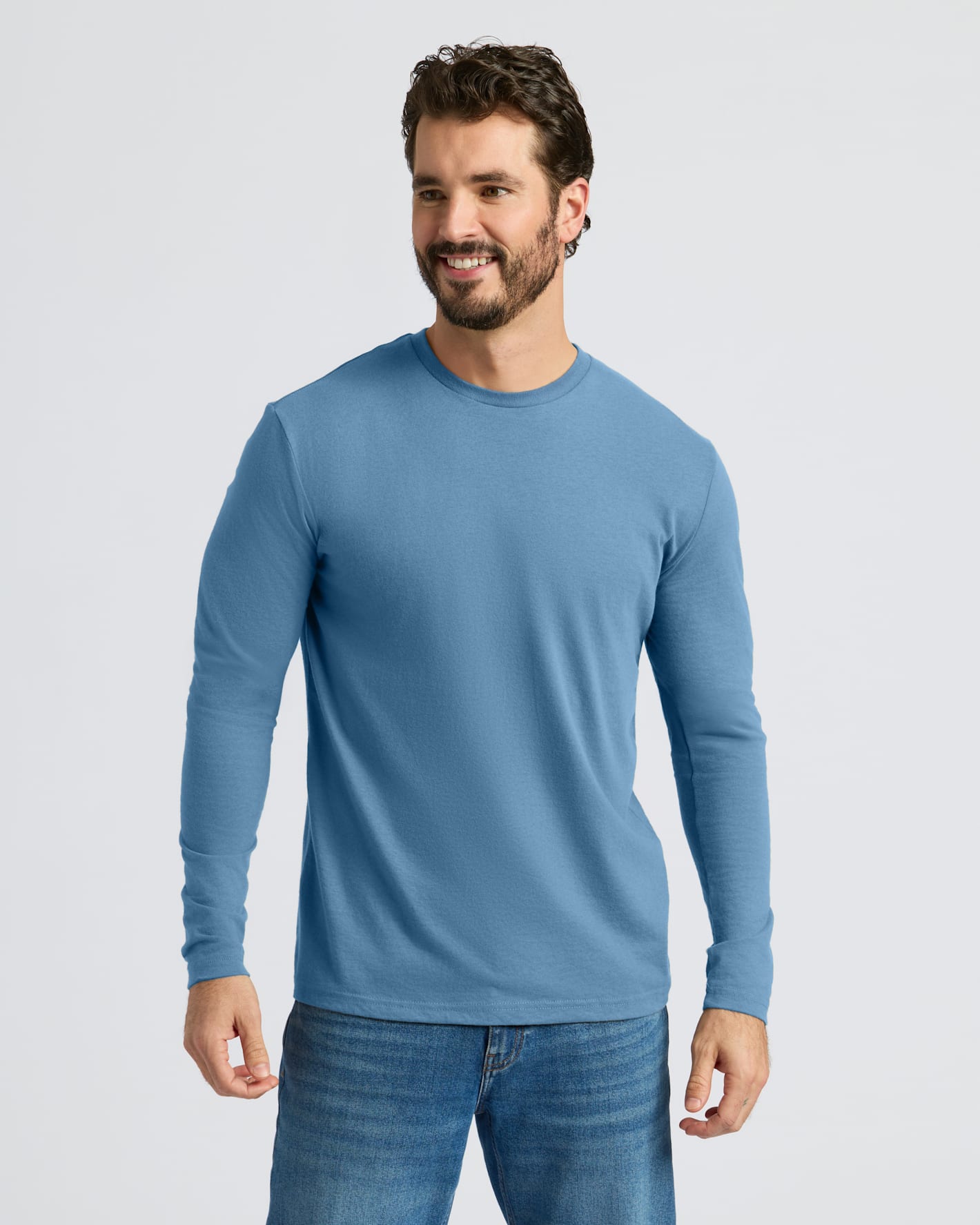 A smiling man wearing a light blue long-sleeve crewneck shirt and blue jeans, standing against a light gray studio background.