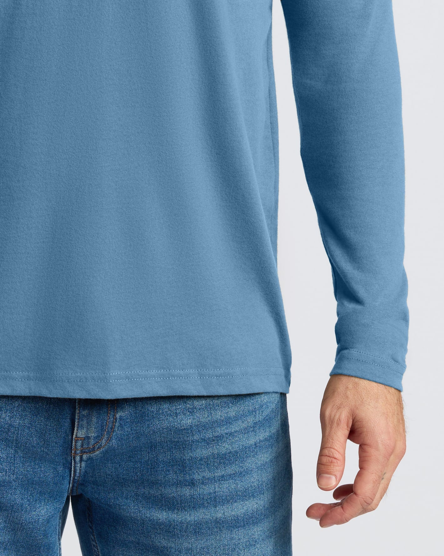 A close-up of a person wearing a light blue long-sleeve t-shirt and blue jeans against a plain light gray background.