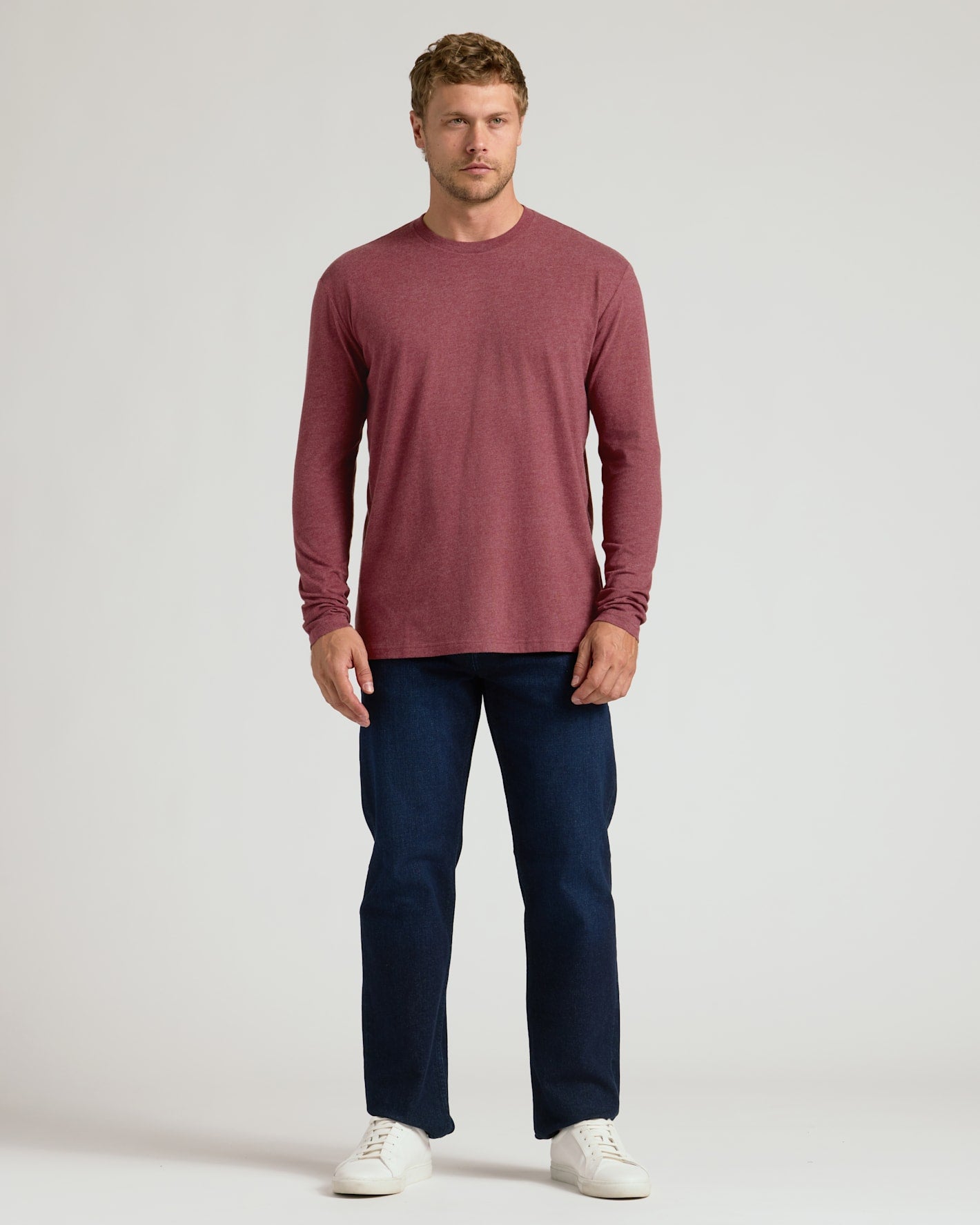 A man wearing a Dark Heather Rustic Apple Classic Long Sleeve Crew Neck shirt paired with dark jeans and white sneakers, standing against a plain light background.