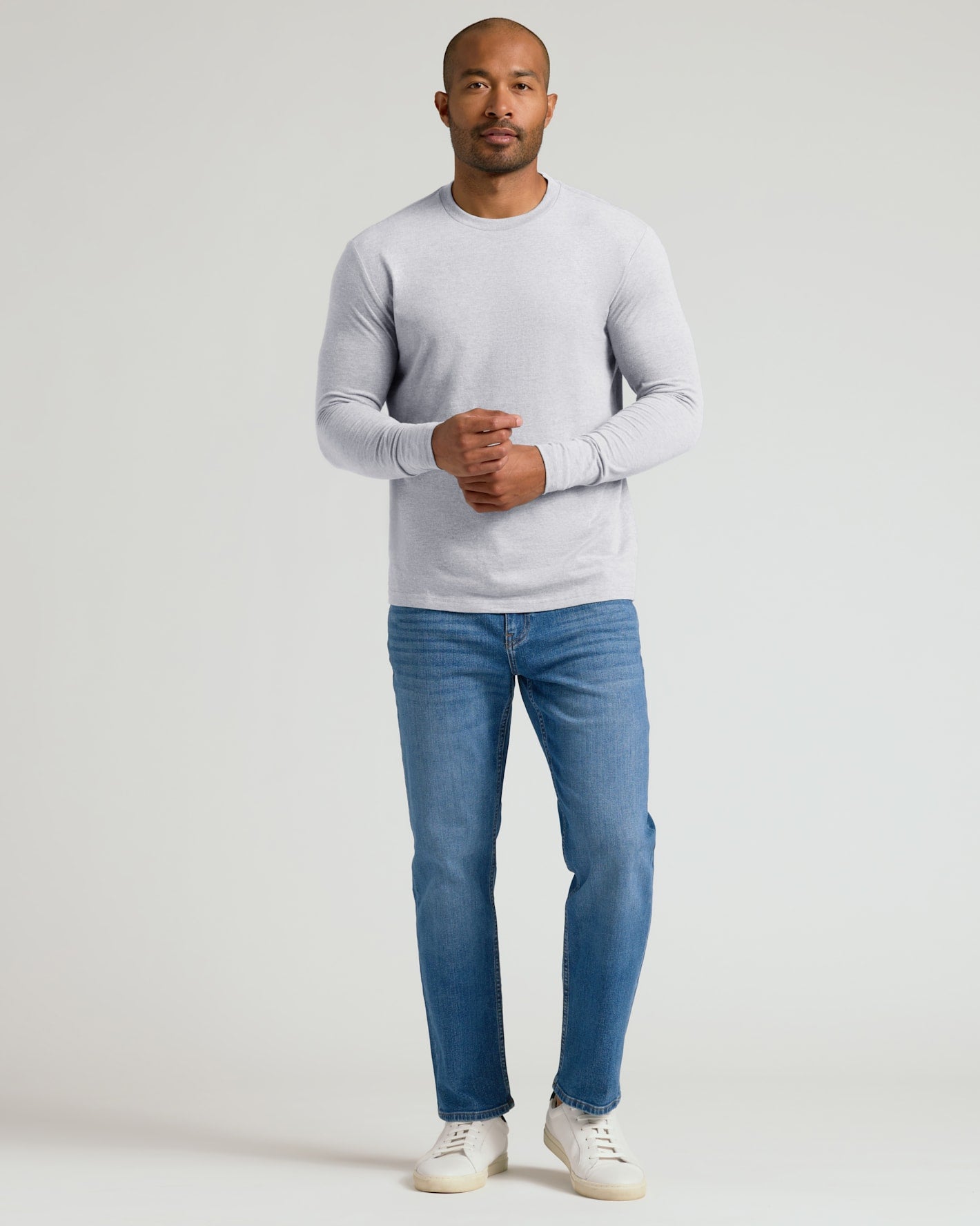 A man wearing a light gray long-sleeve crewneck sweater, blue jeans, and white sneakers, standing against a plain light gray studio background.