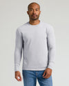 A man wearing a light gray long-sleeve crewneck shirt and blue jeans, standing against a solid light gray background.