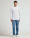 A man modeling a white long-sleeve crewneck t-shirt paired with blue jeans and white sneakers, standing against a light gray studio background.