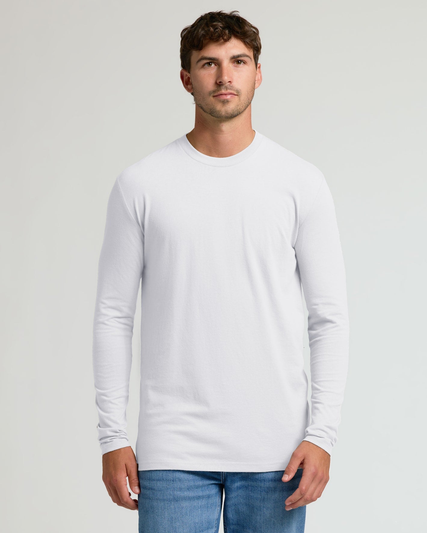A man wearing a plain white long-sleeve crewneck t-shirt and blue jeans, standing against a light gray studio background.