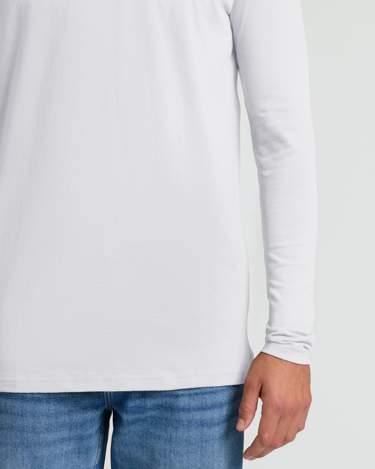 A close-up of a person wearing a plain white long-sleeve top and blue jeans against a light gray studio background.