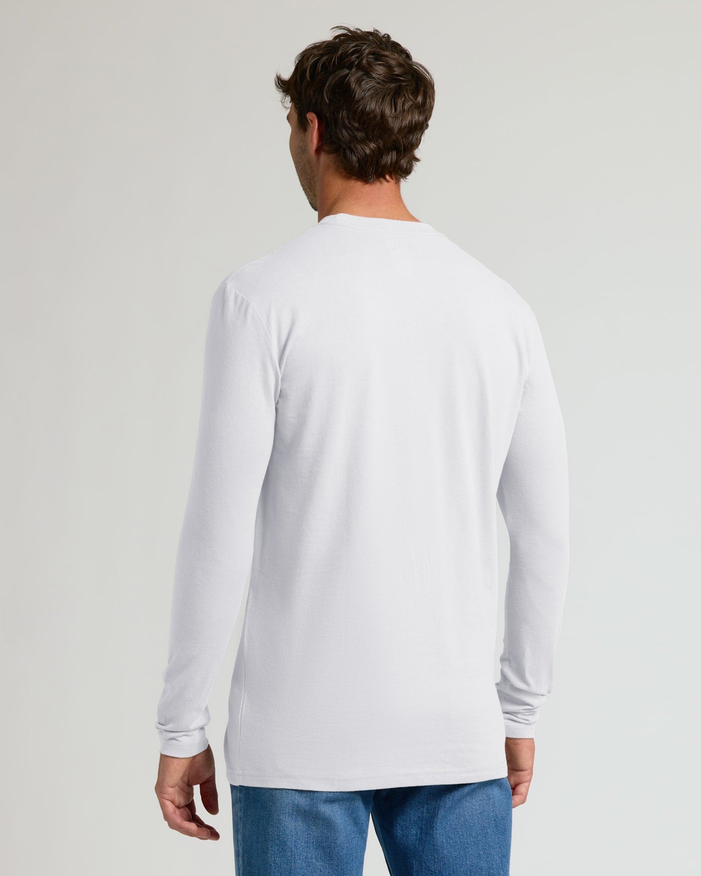 A man viewed from the back wearing a plain white long-sleeve crewneck shirt and blue jeans, standing against a light gray studio background.