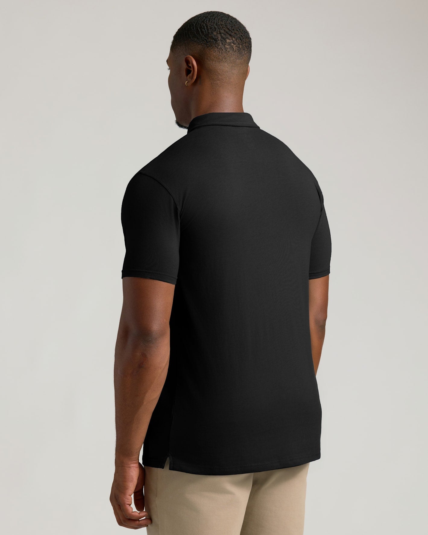 A man viewed from the back wearing a black short-sleeve polo shirt and beige trousers, photographed against a plain light gray studio background.