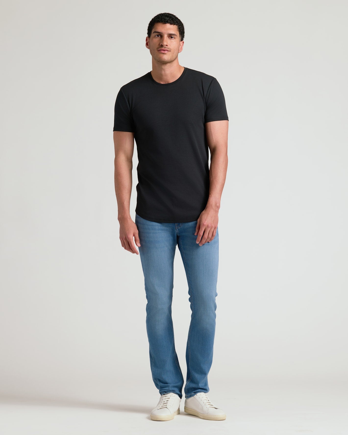 A man wearing a black Tall Pima Cotton Blend Curved Hem Crew 4-Pack t-shirt paired with light blue jeans and white sneakers, standing against a plain background.