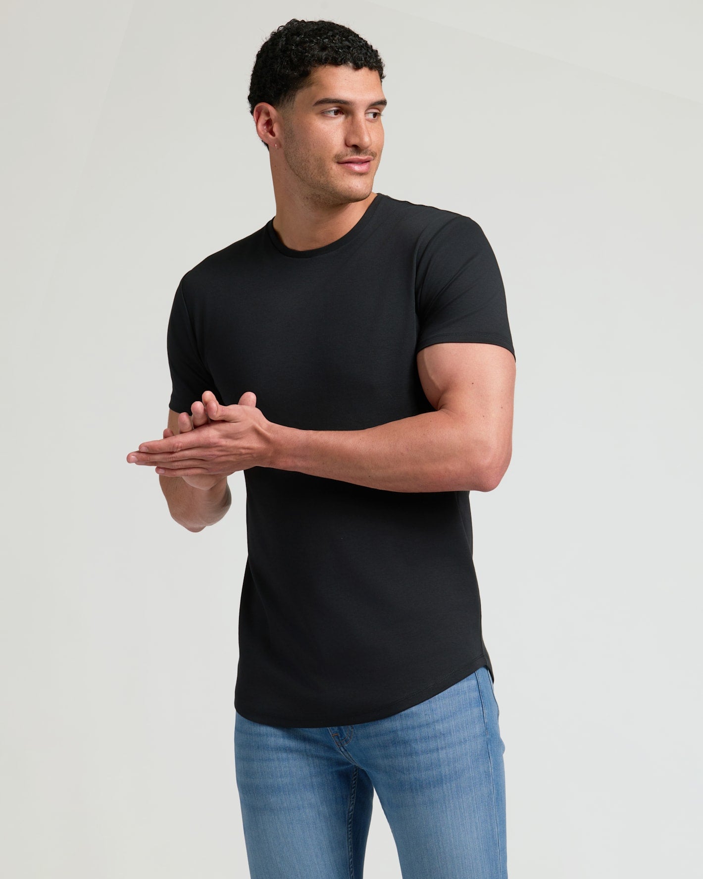 Man wearing a black Tall Pima Cotton Blend Curved Hem Crew 4-Pack shirt paired with light blue jeans against a neutral background, showcasing a modern men's basics style.