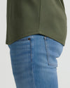 Side view of a person wearing light blue jeans and an olive green Grove Tall Pima Cotton Blend Curved Hem Crew, highlighting the curved hem detail against a plain background.