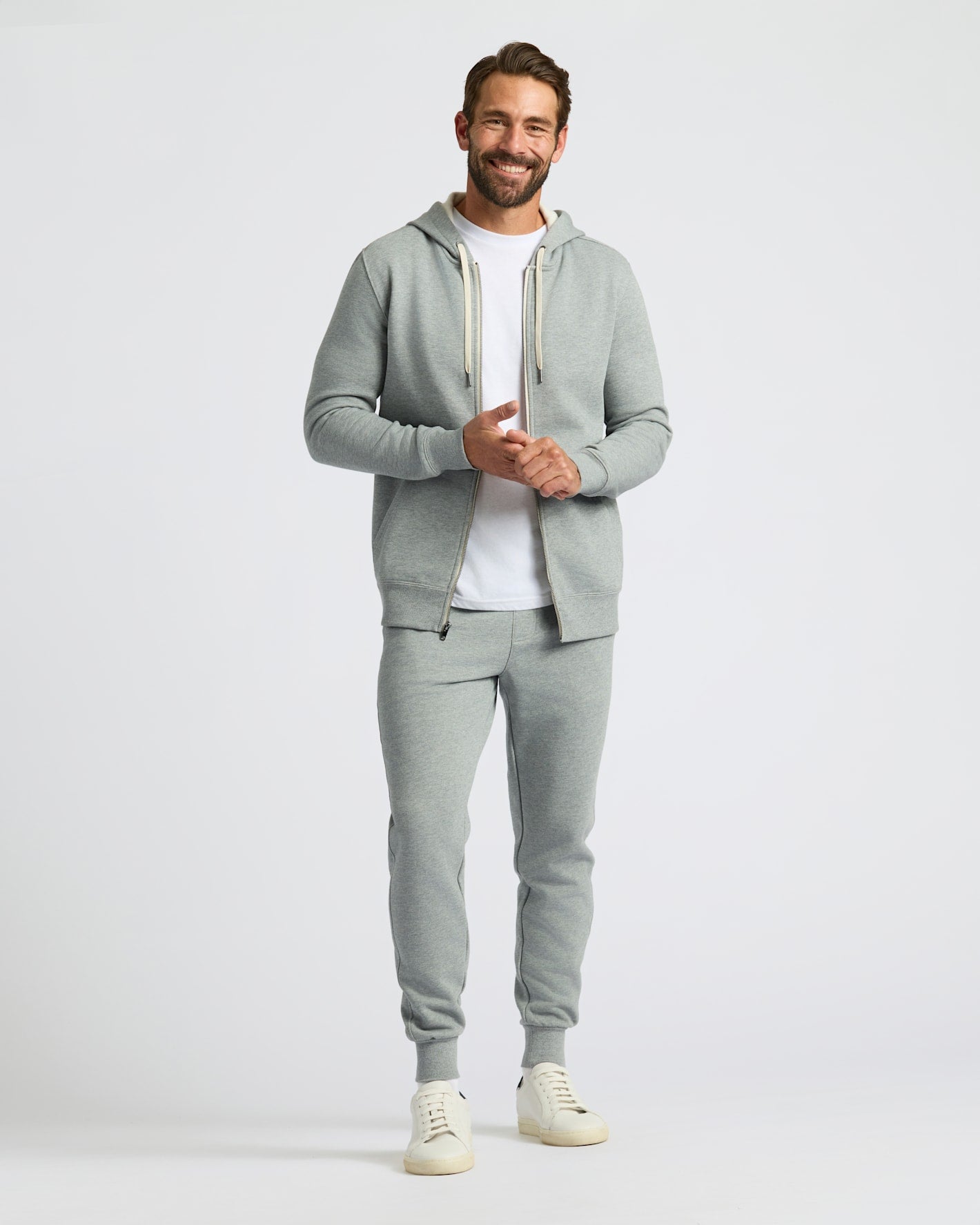 A man modeling a light gray two-piece tracksuit featuring a zip-up hoodie with white drawstrings and matching jogger pants, styled over a white t-shirt and cream sneakers against a light gray studio background.