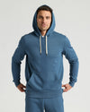 Man wearing a blue Fiord Fleece French Terry Pullover Hoodie with white drawstrings, posing with hands resting on hips against a neutral background.