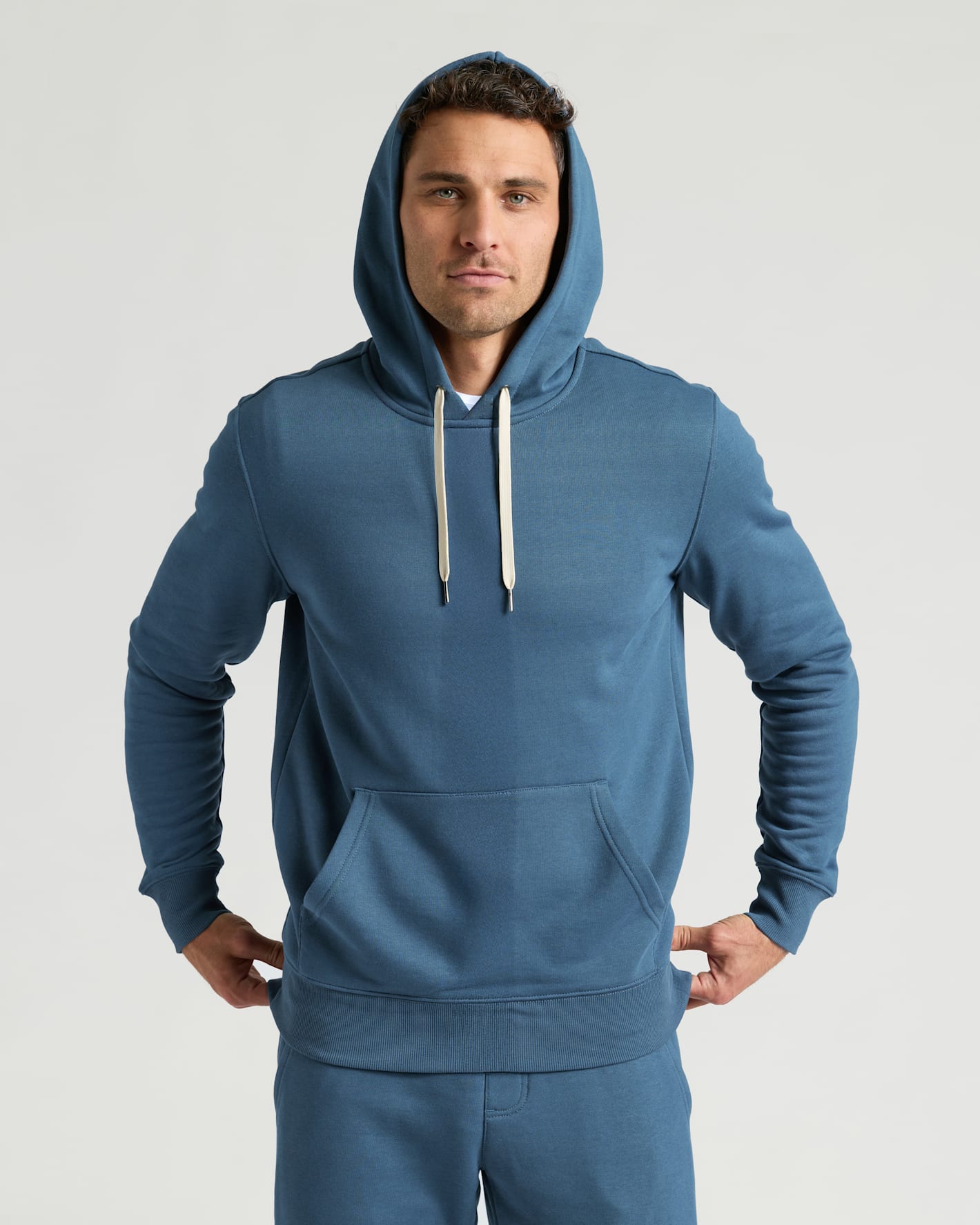 Man wearing a blue Fiord Fleece French Terry Pullover Hoodie with white drawstrings, posing with hands resting on hips against a neutral background.