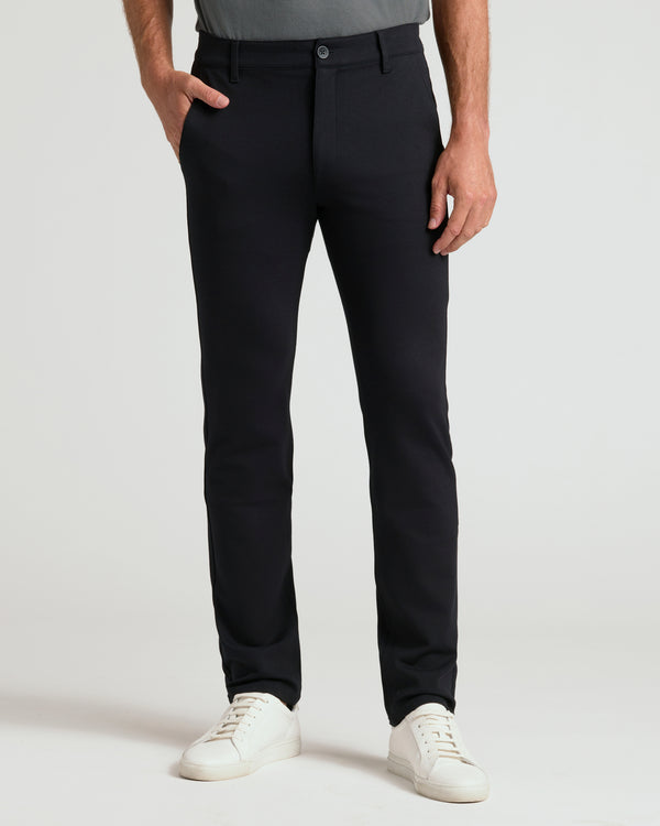 A person wearing slim-fit black trousers, paired with a gray top and white sneakers, standing against a light gray studio background.