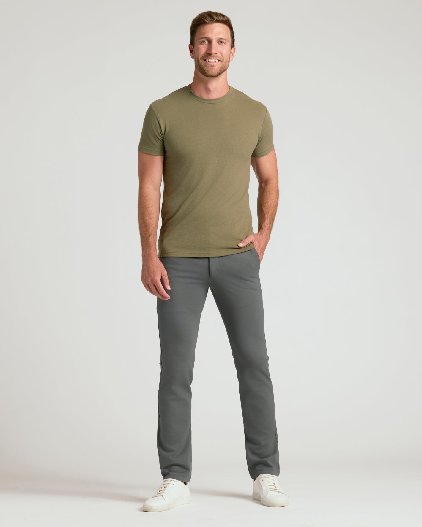 A man modeling a short-sleeve crewneck t-shirt in olive green and gray trousers, standing against a light gray studio background.