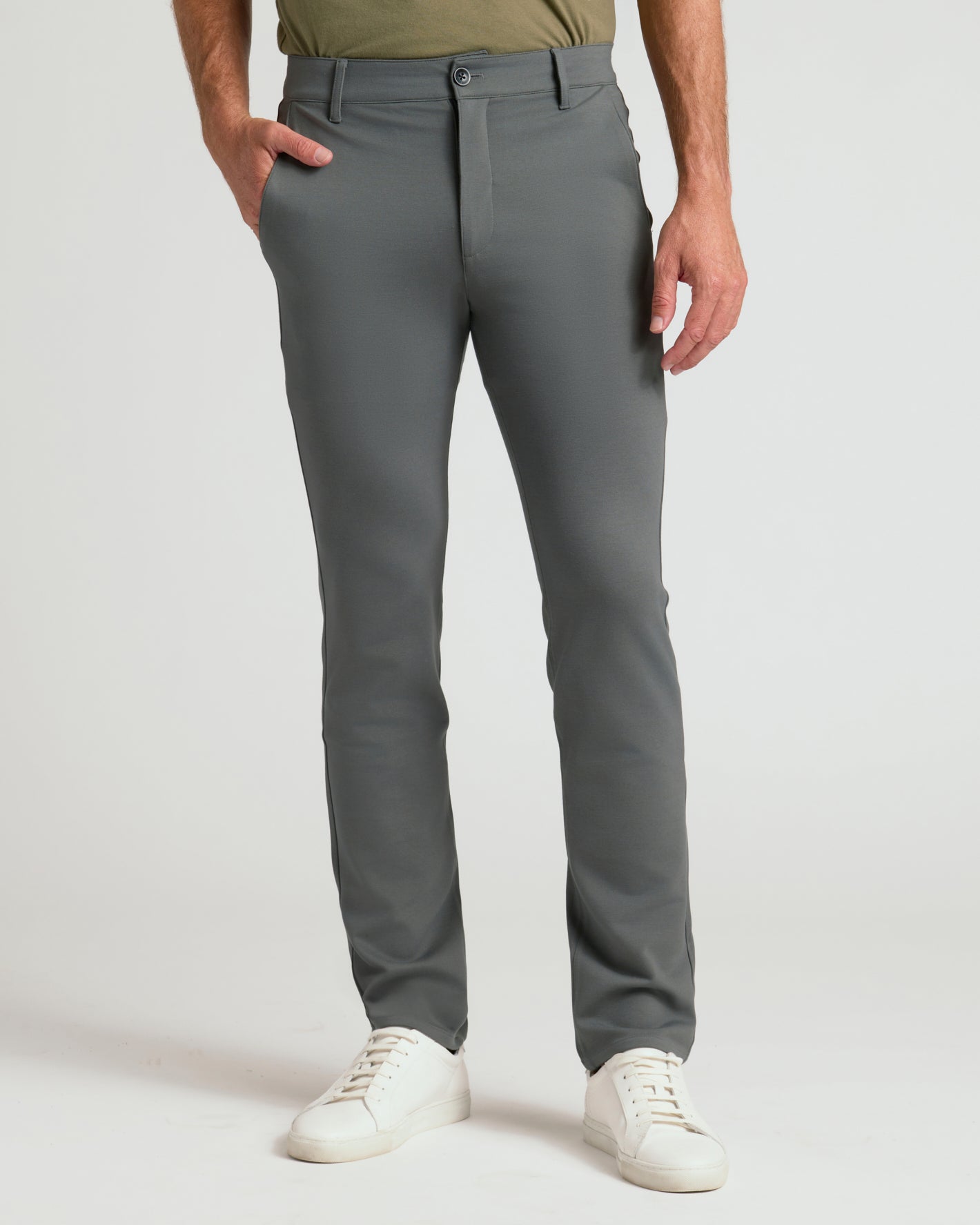 A man wearing slim-fit gray dress pants with an olive green shirt and white sneakers, shown from the waist down against a light gray studio background.