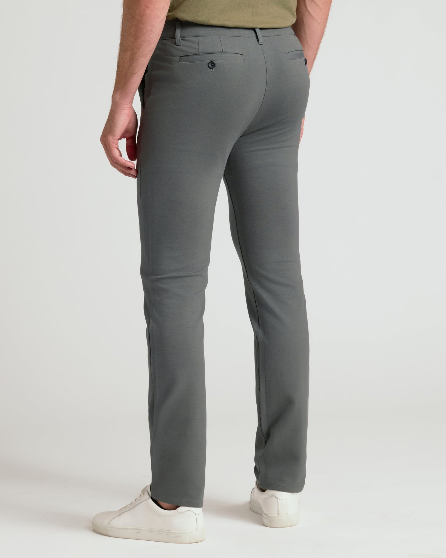 A person shown from the back wearing slim-fit gray trousers, a muted olive green top, and white sneakers against a clean light gray studio background.