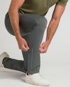 A man wearing slim-fit gray trousers and white sneakers, kneeling down to adjust or pull the side of his left pant leg. He is wearing an olive green t-shirt against a plain light gray studio background.