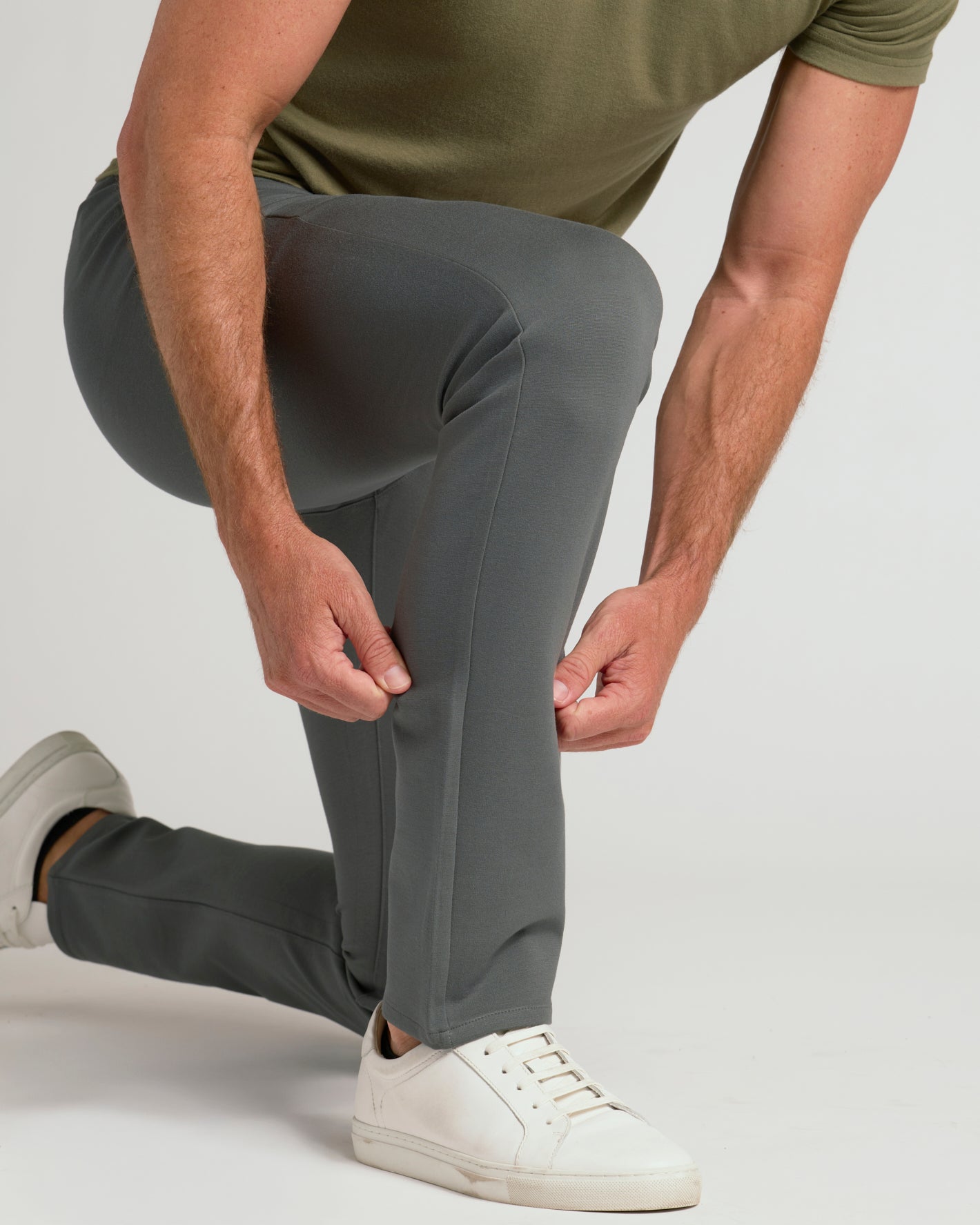 A man wearing slim-fit gray trousers and white sneakers, kneeling down to adjust or pull the side of his left pant leg. He is wearing an olive green t-shirt against a plain light gray studio background.