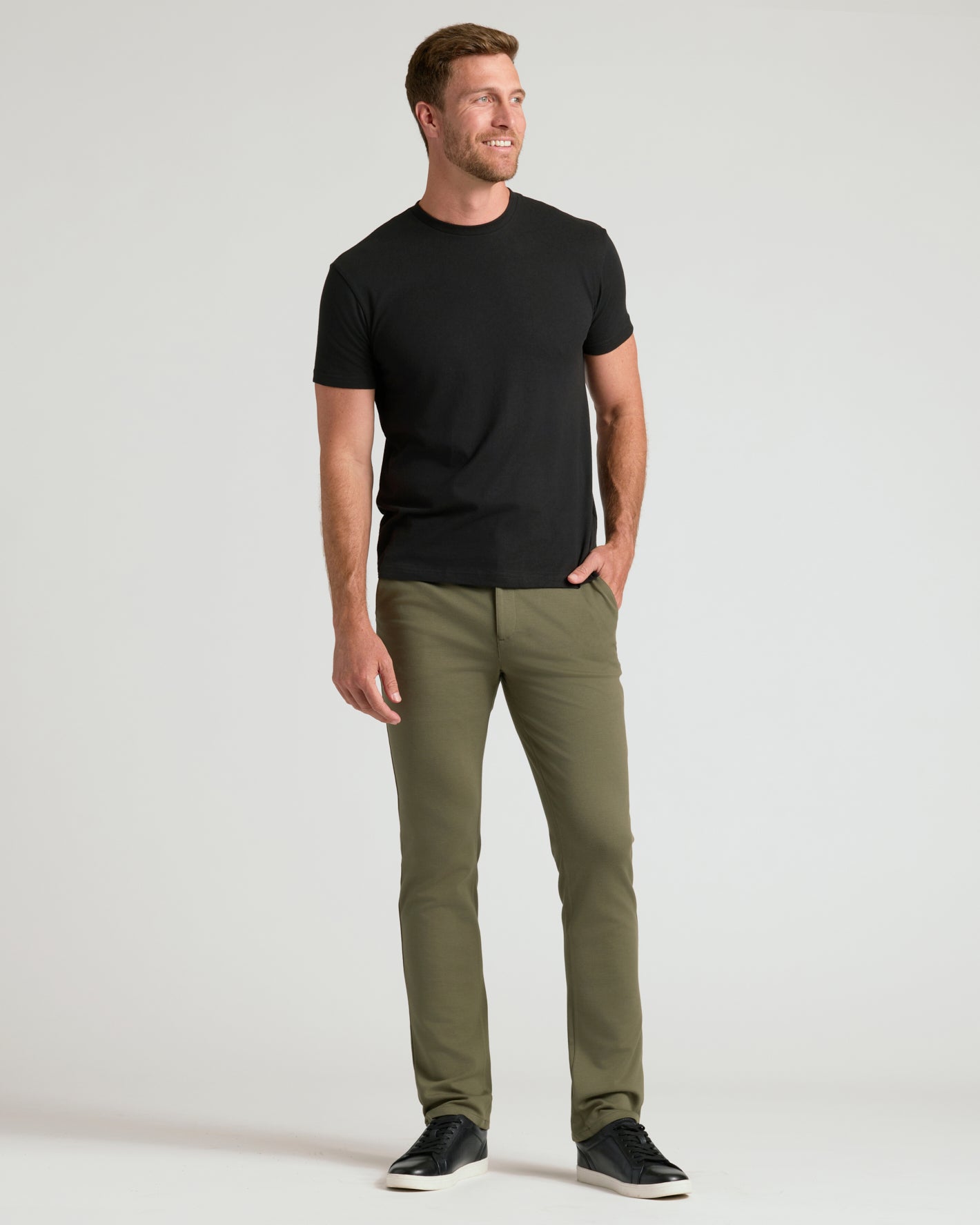 A man modeling a black short-sleeve crewneck t-shirt and olive green slim-fit trousers, paired with black sneakers, against a light gray studio background.