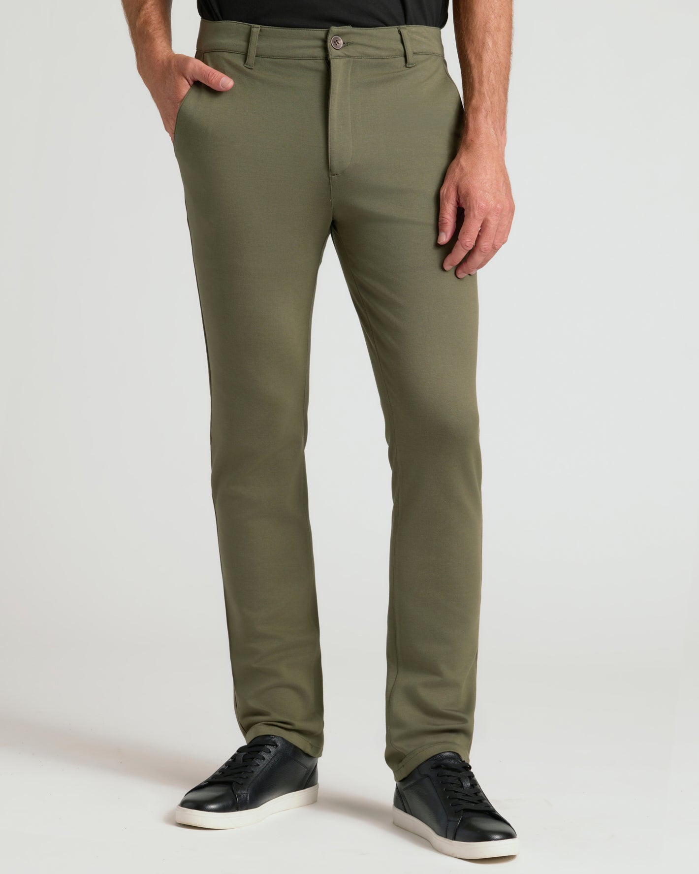 A person wearing olive green slim-fit chinos with black sneakers, shown from the waist down against a light gray studio background.