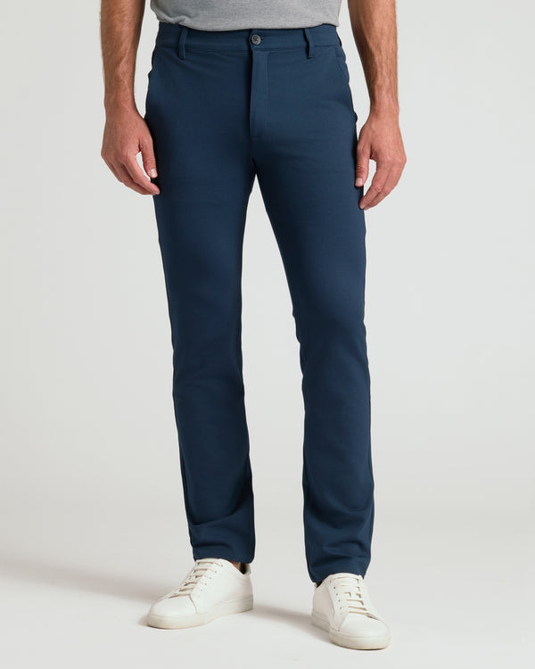A person modeling a pair of slim-fit navy blue trousers, paired with a gray t-shirt and white sneakers, against a plain light gray studio background.