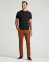 A man modeling a black short-sleeve crewneck t-shirt and rust-brown slim-fit trousers, paired with black sneakers, against a light gray studio background.