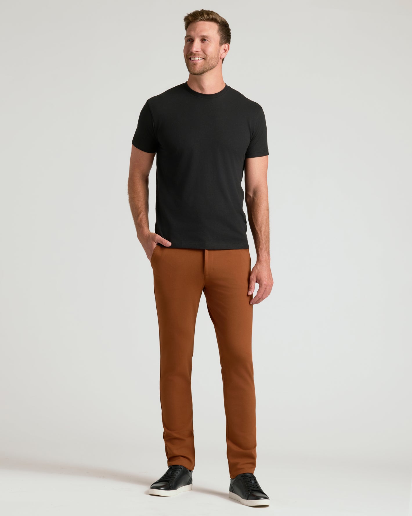 A man modeling a black short-sleeve crewneck t-shirt and rust-brown slim-fit trousers, paired with black sneakers, against a light gray studio background.