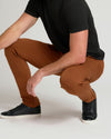 A man crouching in a studio setting, wearing slim-fit rust-brown pants, a black t-shirt, and black sneakers with white soles.