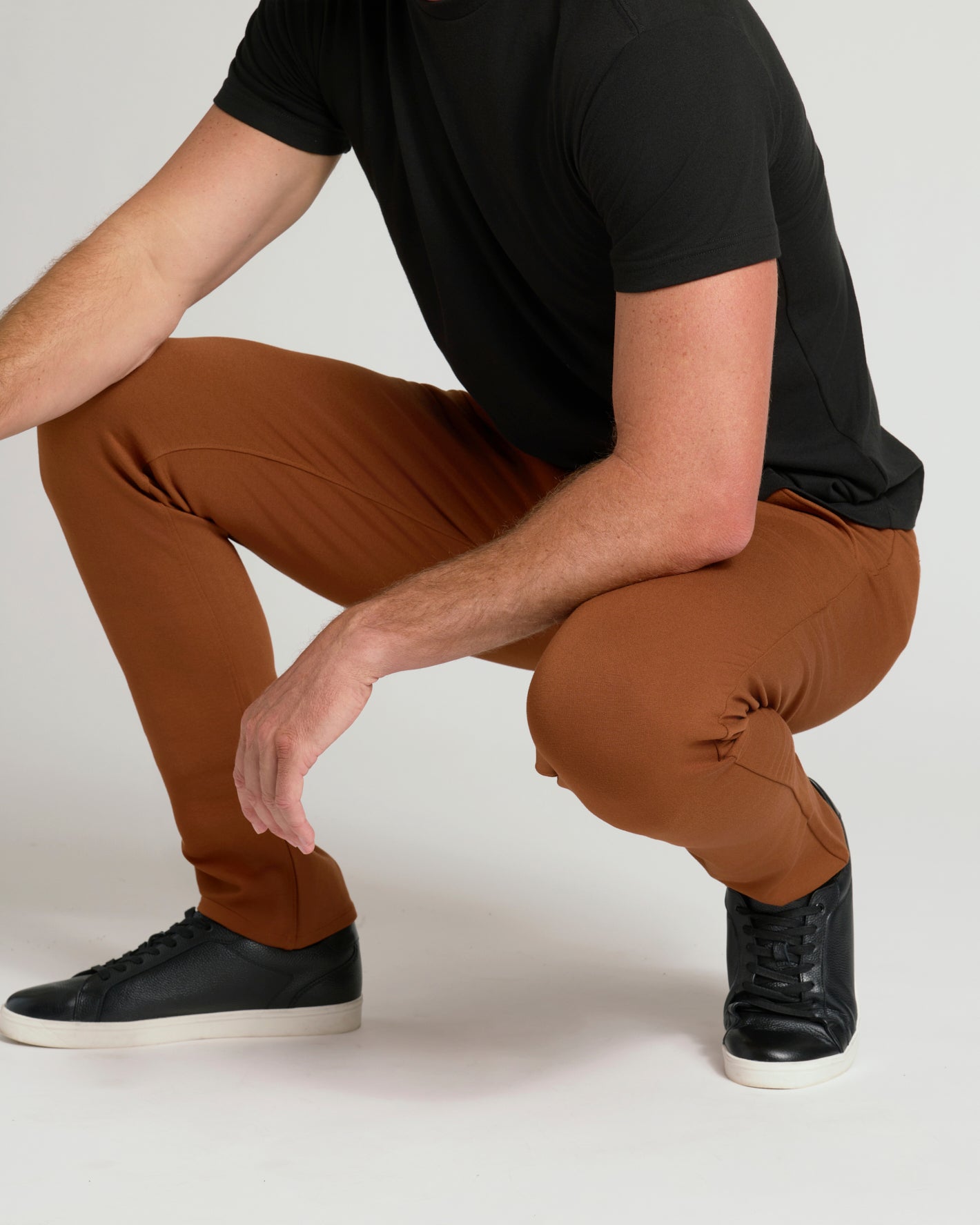 A man crouching in a studio setting, wearing slim-fit rust-brown pants, a black t-shirt, and black sneakers with white soles.