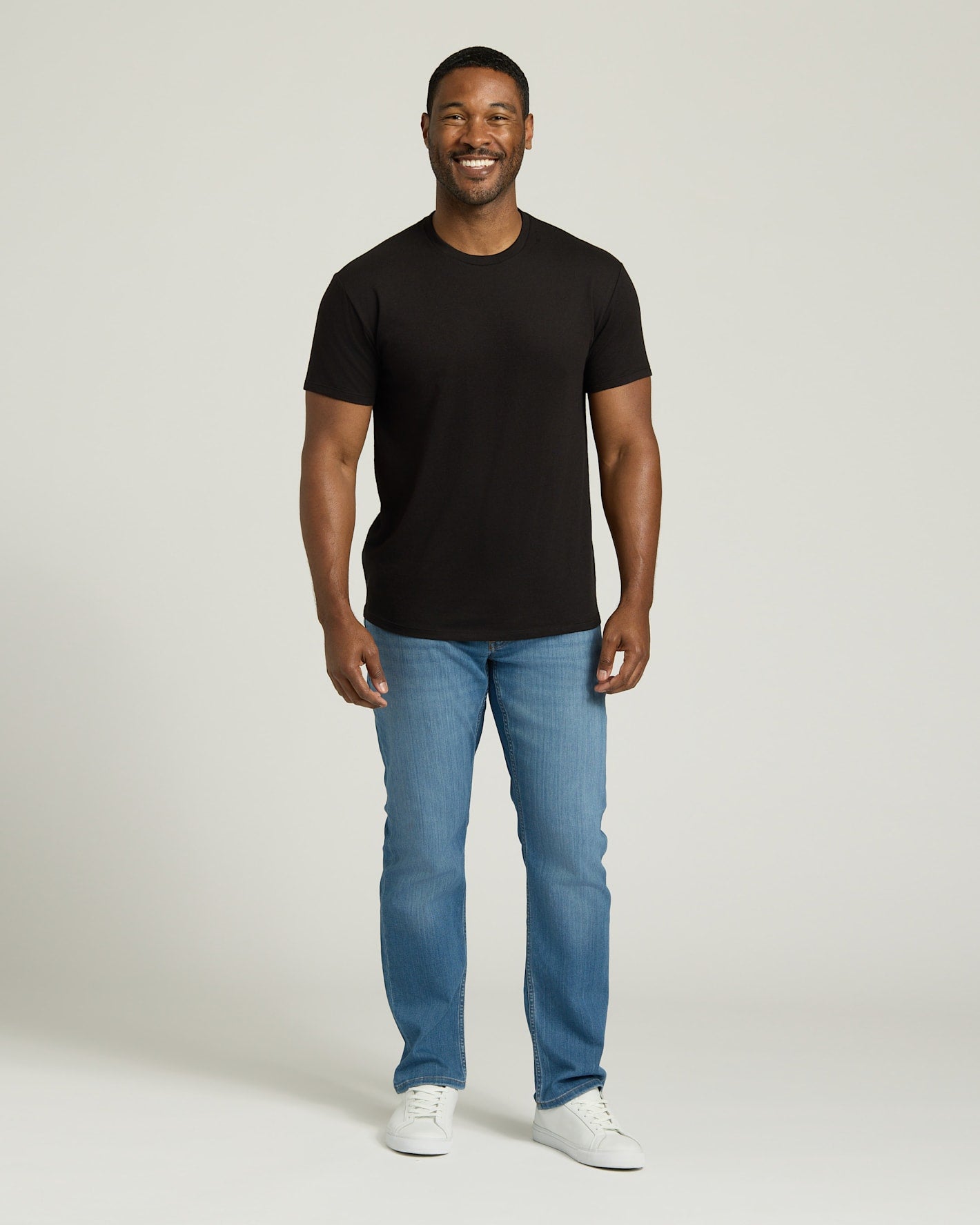 Man wearing a black t-shirt, blue jeans, and white sneakers on a plain background. MODEL_INFO: Model is 5'8" wearing size M short