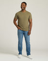 Man wearing a green t-shirt and blue jeans standing against a plain background. MODEL_INFO: Model is 5'8" wearing size M short