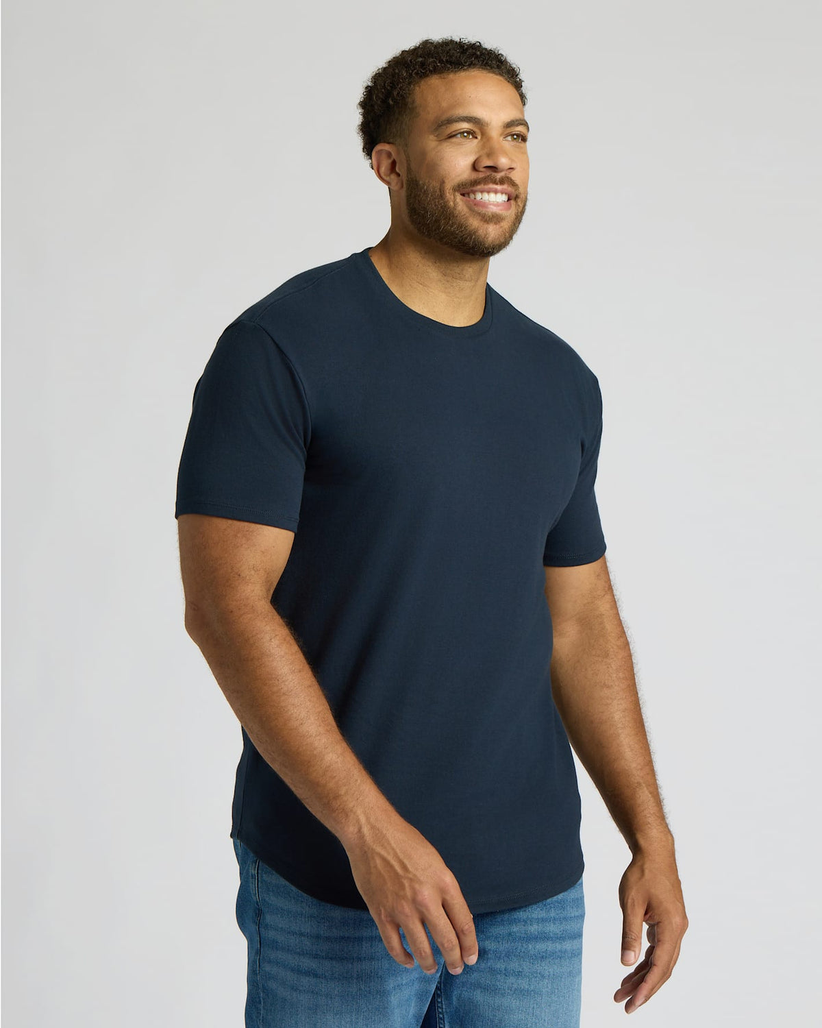 Man wearing a navy blue True Classic Men's Tall Heavyweight Curved Hem Crew – Premium Heavyweight Cotton Blend, Extended Length, Athletic Fit, Structured Everyday T-Shirt with jeans, smiling and looking to the side.