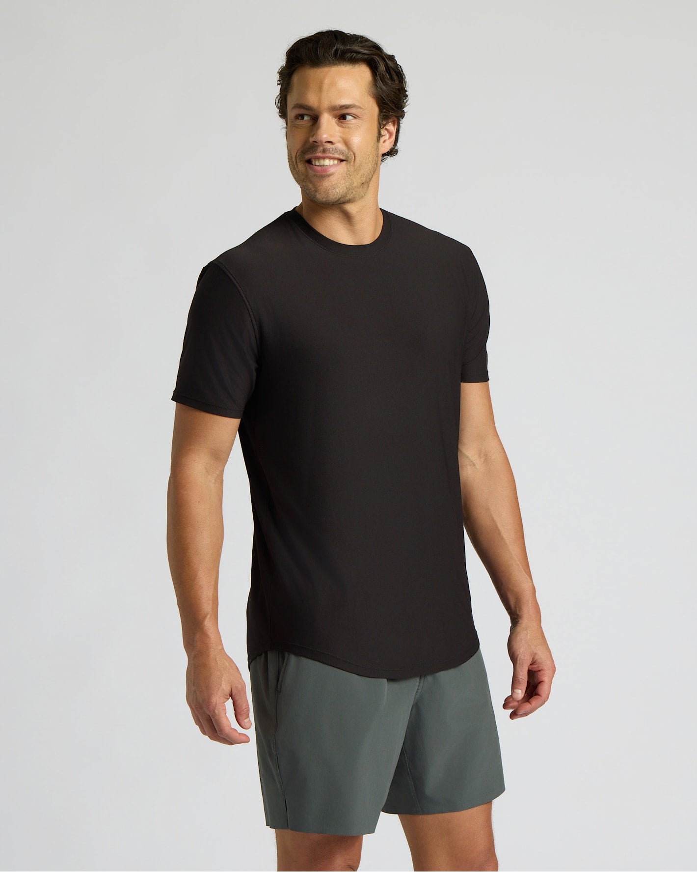 Man wearing a black Active Curved Hem Crew Tee - Men's Moisture Wicking Performance T-Shirt with curved hem and short sleeves, paired with gray shorts against a plain background.