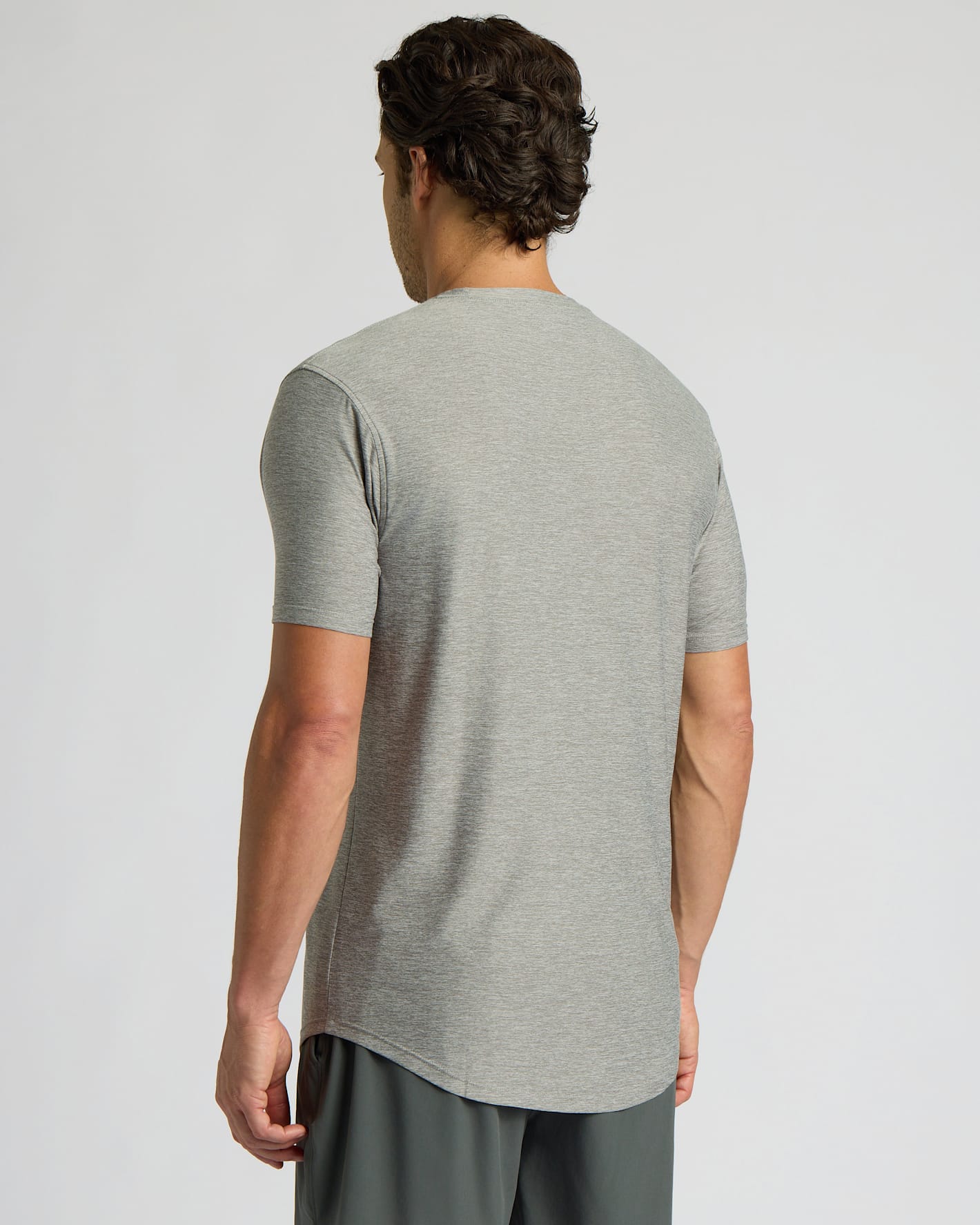 A man viewed from the back wearing a light gray short-sleeve t-shirt and dark gray athletic pants, set against a plain light gray studio background.