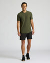 A man modeling a short-sleeve crewneck athletic t-shirt in olive green, paired with black athletic shorts and black running shoes, against a light gray studio background.