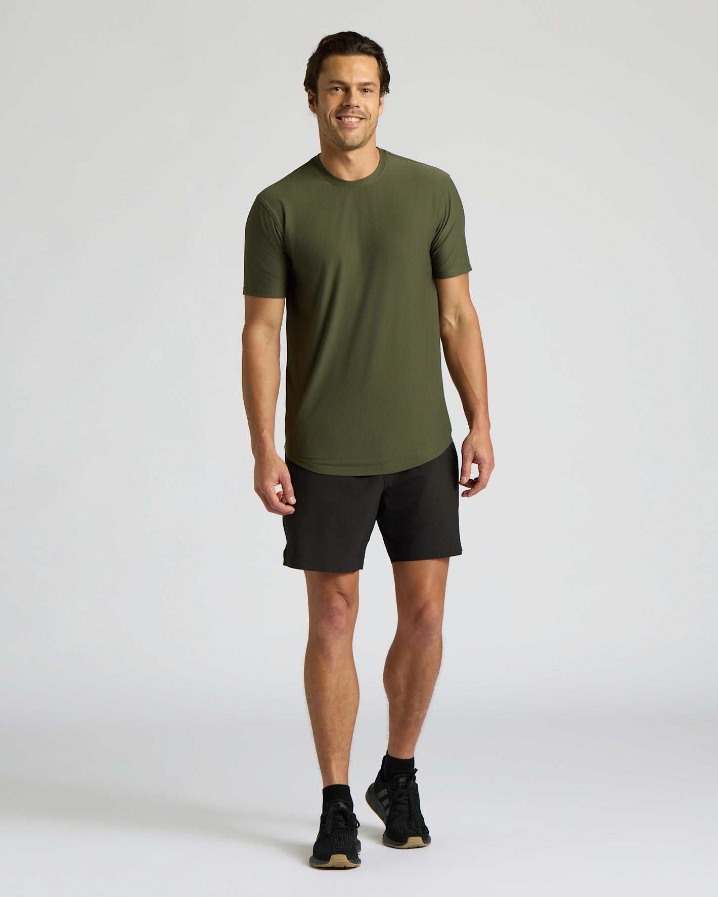 A man modeling a short-sleeve crewneck athletic t-shirt in olive green, paired with black athletic shorts and black running shoes, against a light gray studio background.